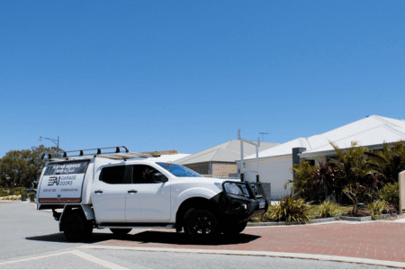 An A1 Garage Doors Vehicle is driving into a driveway, ready to fix a roller door. The vehicle is a white ute, the driveway is red bricks with a cream boarder. The sky is bright blue, it's a sunny day.
